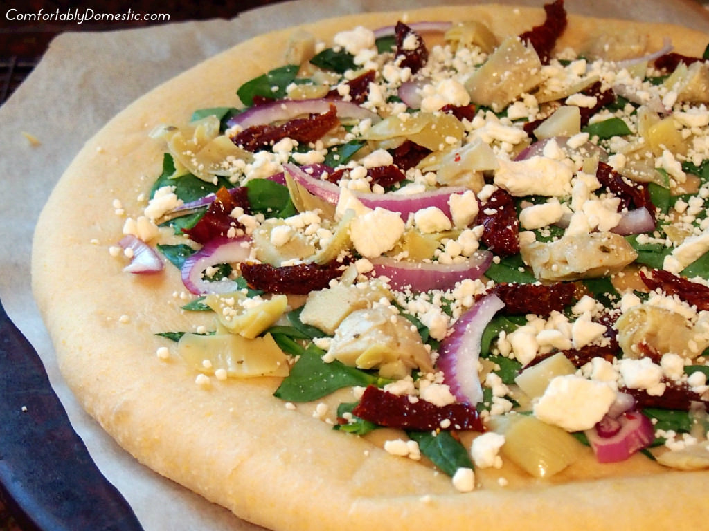 Greek Pizza starts with a tangy tzatziki sauce that unites such Mediterranean flavors as artichoke hearts, sundried tomatoes, spinach, red onion, and a feta cheese into tantalizing, cornmeal-crusted pizza. | ComfortablyDomestic.com