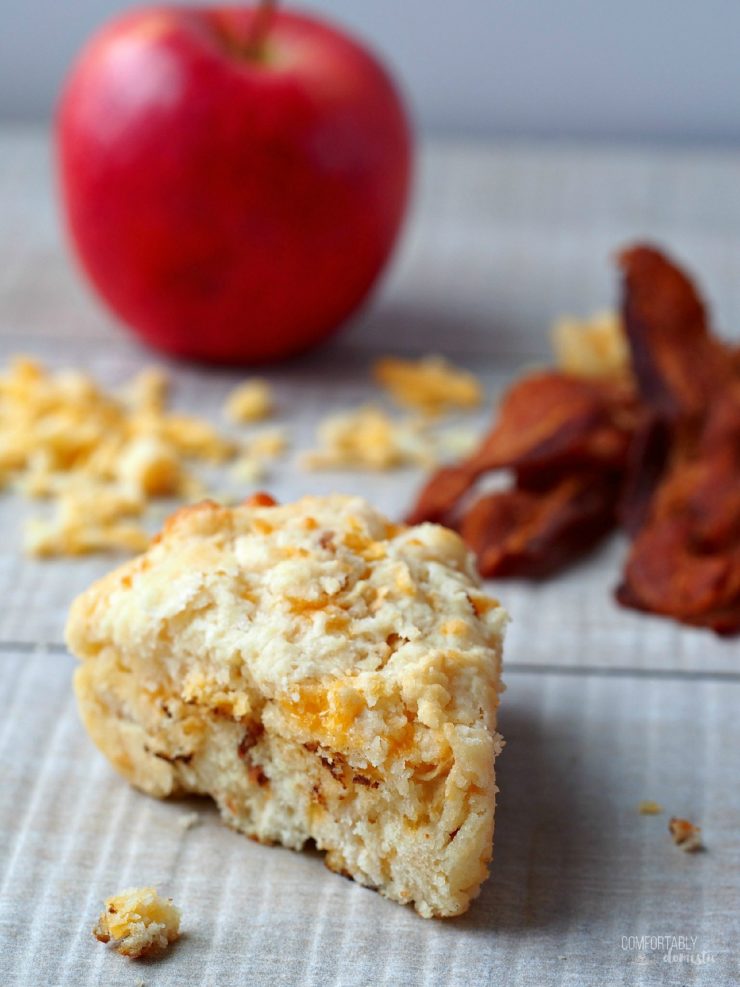 A single Apple Cheddar Bacon Scone with shredded cheese, crisp bacon, and an apple in the background.