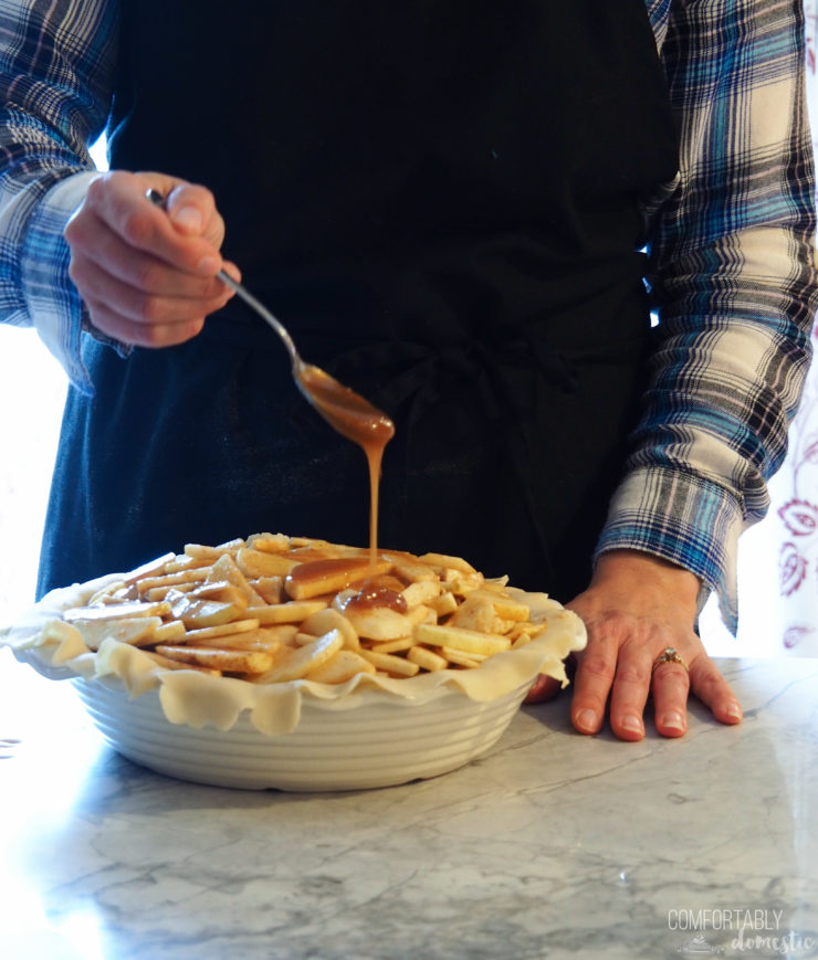 a hand drizzling caramel over the apples in the filling of a caramel apple pie.