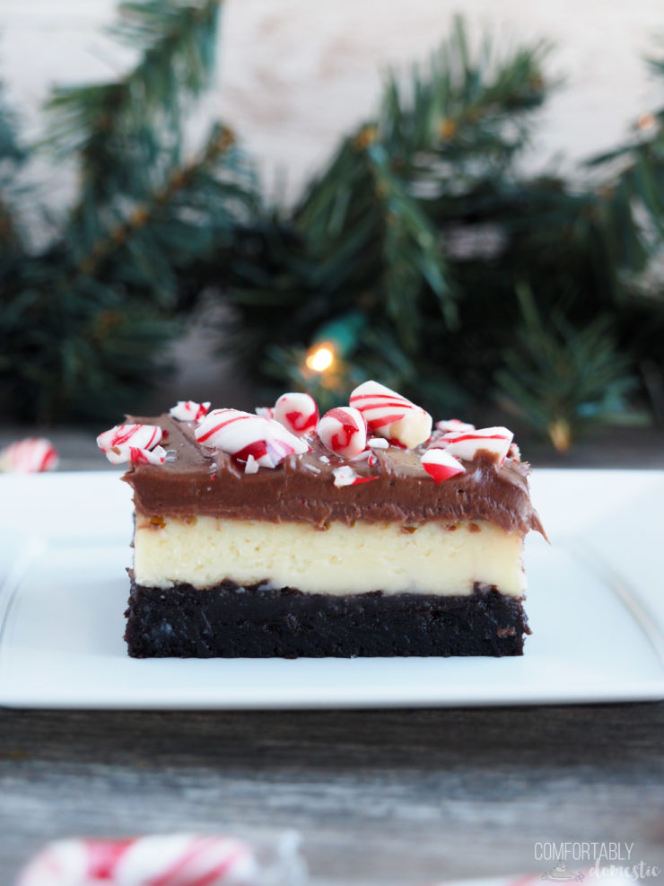 Close up photo of a layered peppermint cheesecake brownie on a white plate with pine garland in the background.