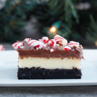 Close up of a layered peppermint cheesecake brownie on a white plate with pine garland and twinkle lights in the background.