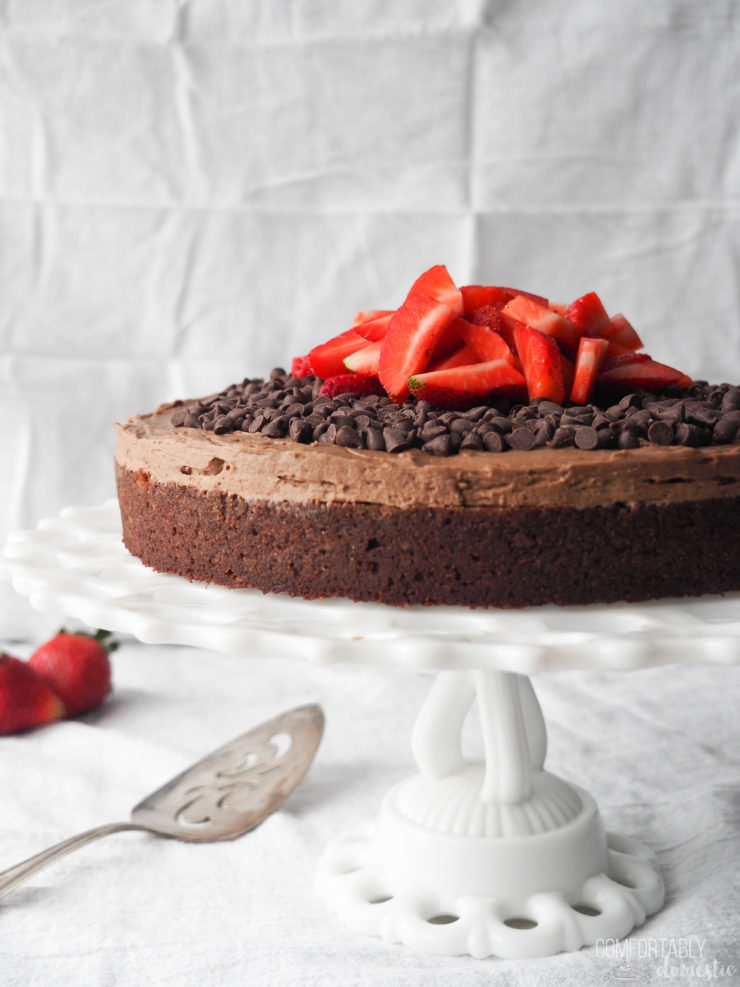 Gluten-free-fudge-cake-frosted-with-chocolate-chips-and-strawberries on a white pedestal plate.