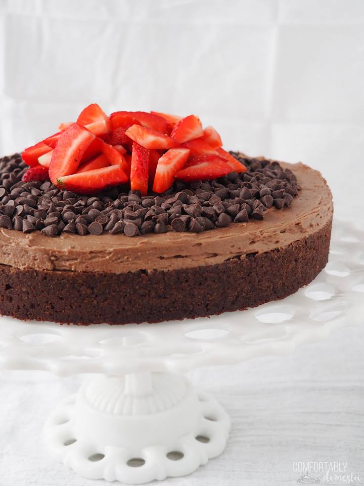 Gluten free chocolate cake with chocolate frosting, topped with mini chocolate chips and sliced fresh strawberries sitting on a white cake pedestal plate. 