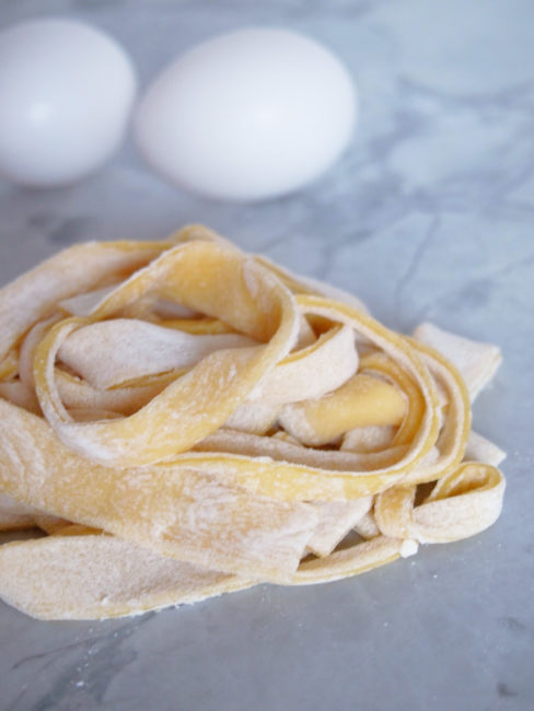 a pile of homemade egg noodles on white marble with two eggs in the background.