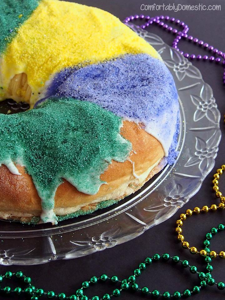 close up photo of Mardi Gras King Cake covered in green, purple, and yellow sugar and beads in the background.