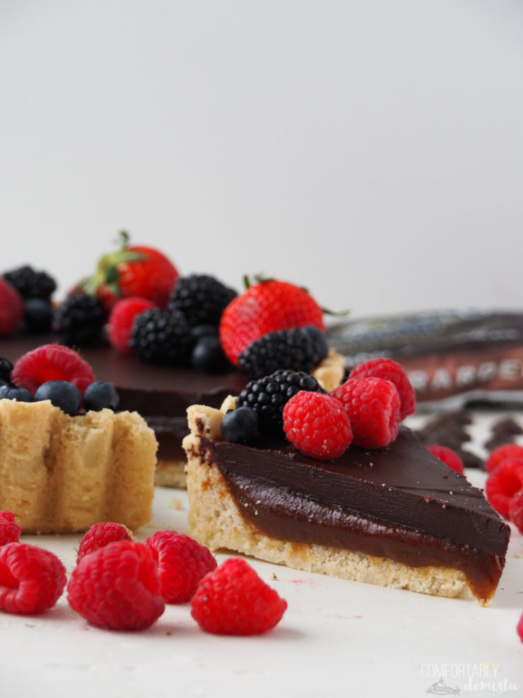 Slice of a Salted Caramel Chocolate Tart with fresh berries on top and scattered around it. 