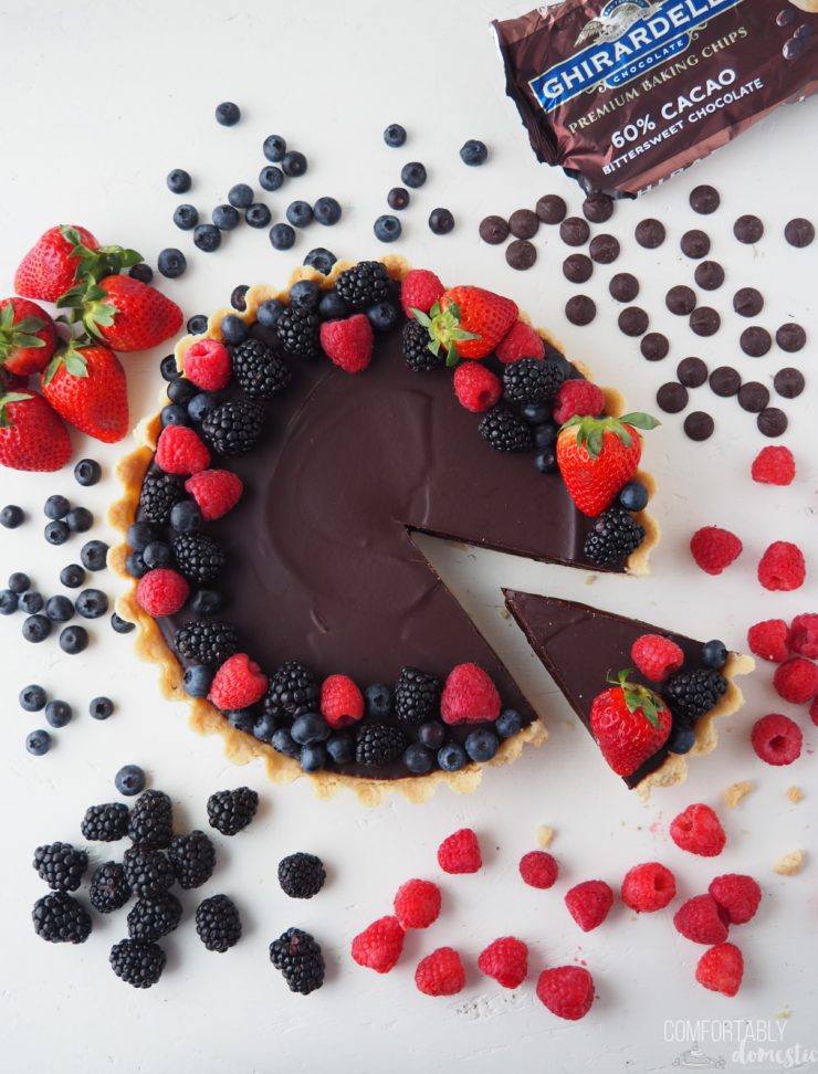 Overhead photo of a Salted Caramel Chocolate Tart on a white background with fresh blueberries, raspberries, strawberries, black berries, and chocolate chips scattered around it.