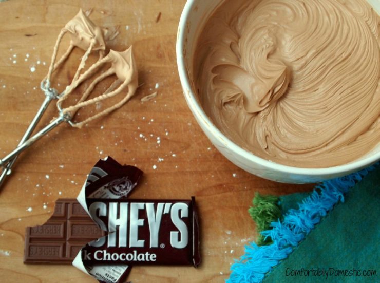 White bowl full of milk chocolate buttercream frosting on a wood board with beaters and a Hershey's chocolate bar.