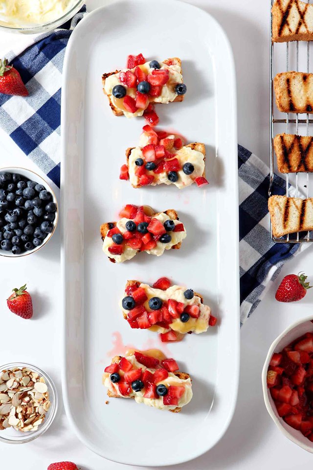 Patriotic Strawberry Bruschetta is strawberry and blueberries atop goat cheese on toast rounds on a white platter.