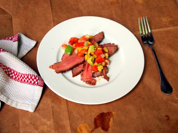 thinly sliced flank steak piled on a white plate with avocado corn salsa on top.