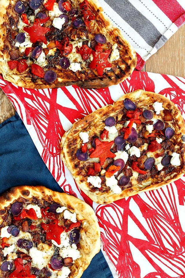 several grilled pizzas with red pepper, blue potatoes, and white goat chees on a red and white tablecloth. 