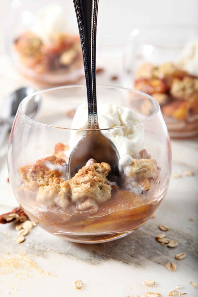 clear stemless wine glasses full of peach cobbler with a spoon on a white background