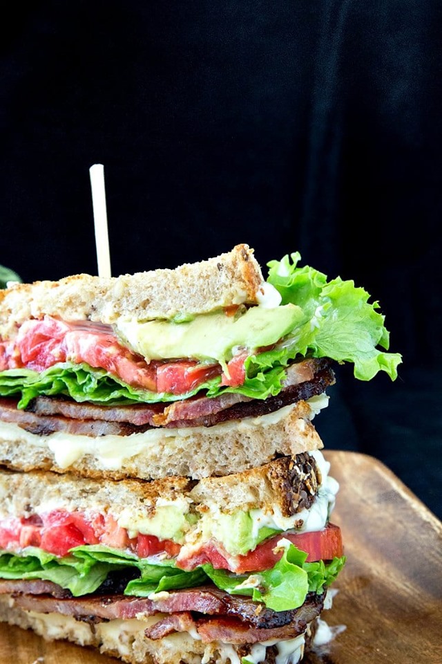 The Best BLT sandwich stacked on a wooden cutting board with a black background. 
