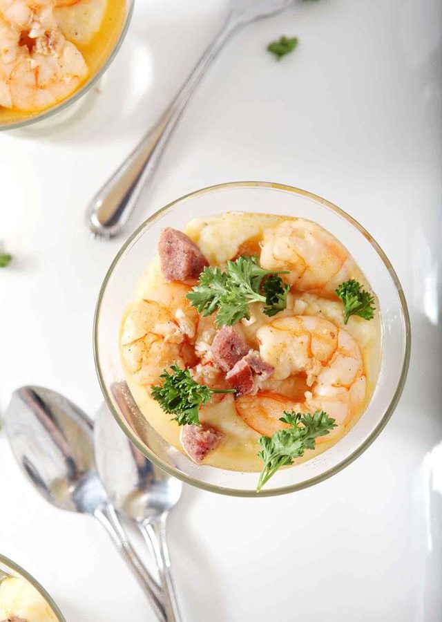 Clear glass bowl of shrimp and grits with herbs scattered over top on a white background.
